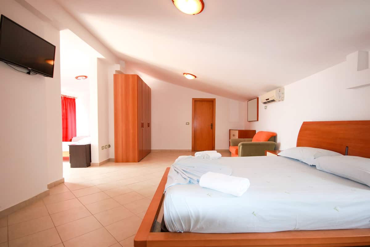 A hotel room with a large bed, a TV mounted on the wall, a wooden wardrobe, a small table with a chair, and a door leading to another room.