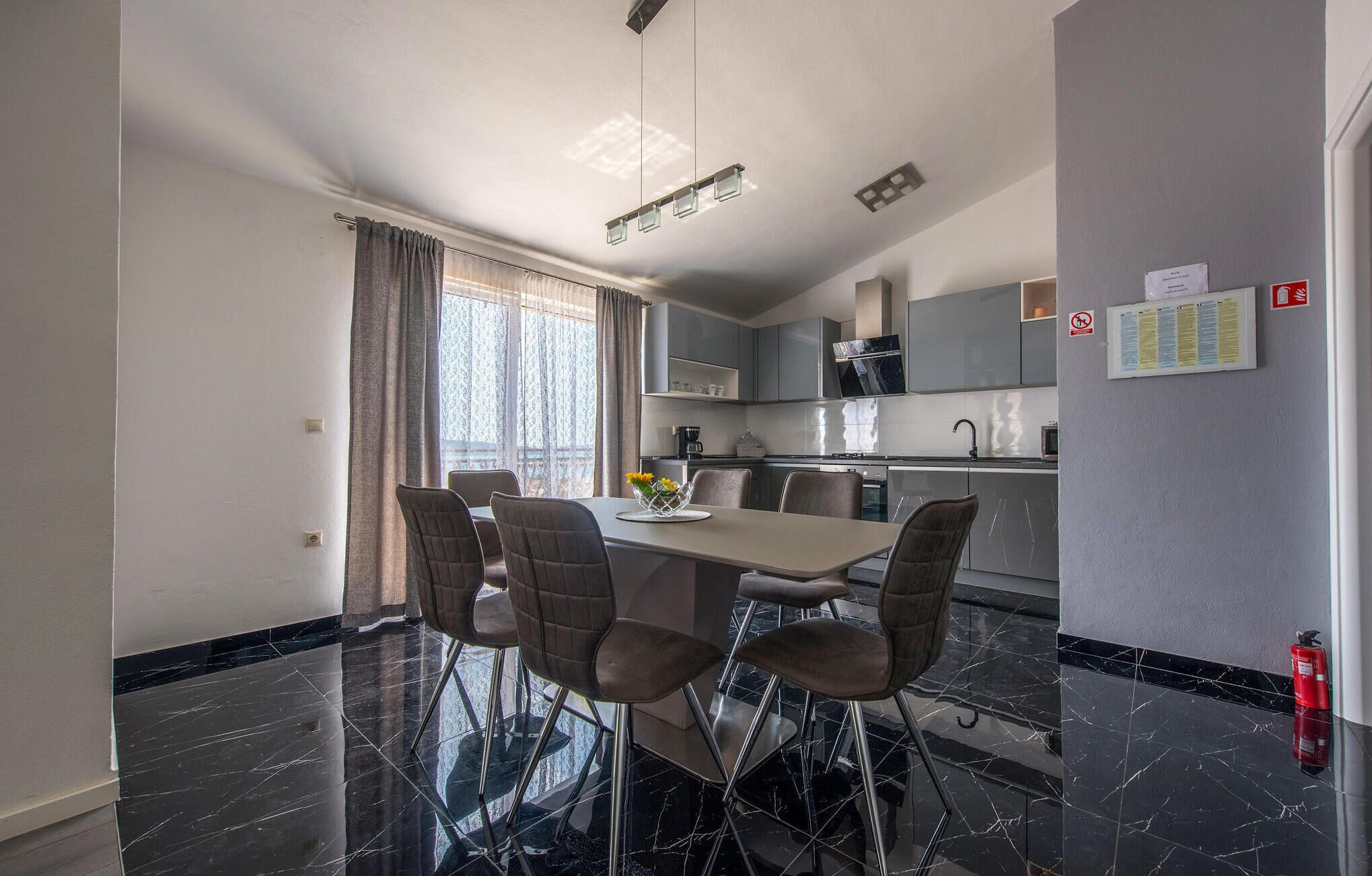 A modern kitchen with a dining area, black marble flooring, and a window with curtains.