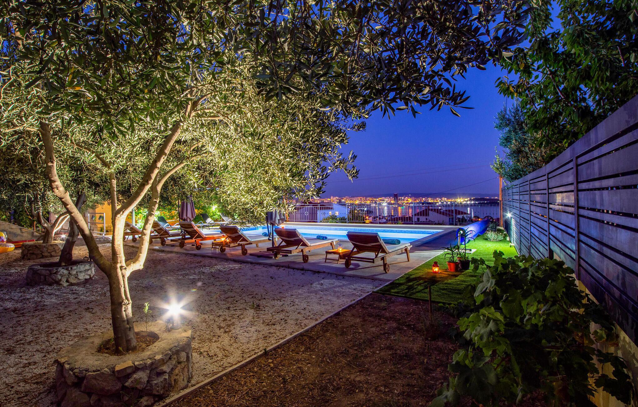 A poolside area with lounge chairs, a small tree, and a view of a cityscape at dusk.