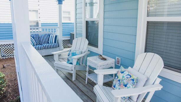 A porch with white Adirondack chairs and a wicker table, blue siding, and a white railing.