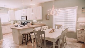 A kitchen with white cabinets, a central island with stools, a chandelier, and a window with blinds.