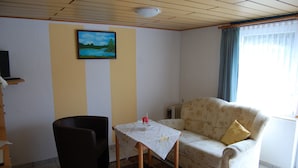 Un salón con sofá, una mesita, una silla, un cuadro en la pared y una ventana con cortinas.