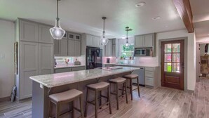A modern kitchen with a large island, bar stools, pendant lighting, and a view of the outdoors through a door. - LD 727 Down River Haus (New Braunfels)