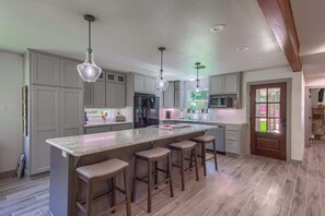 A modern kitchen with a large island, bar stools, pendant lighting, and a view of the outdoors through a door.