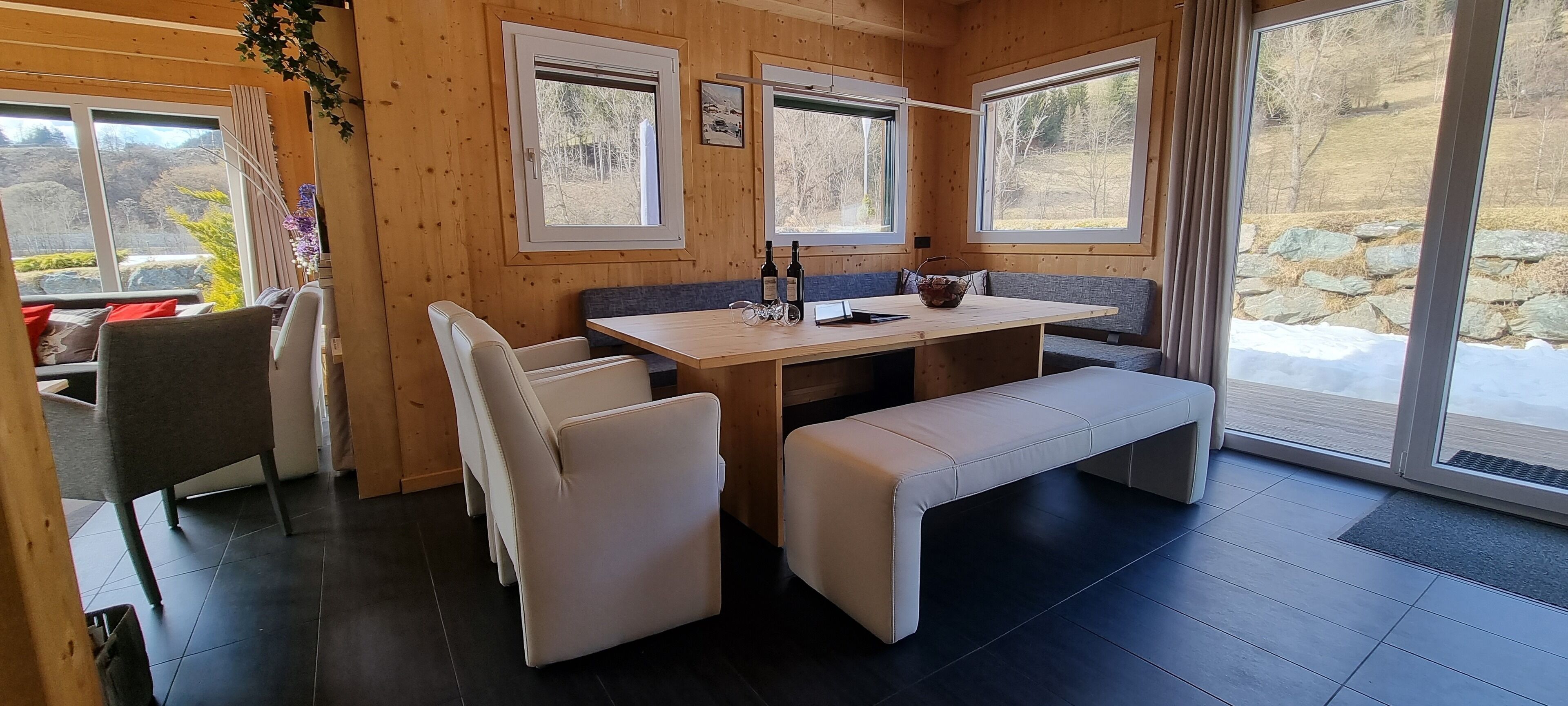 L’intérieur chaleureux d’un chalet, avec un décor en bois, une table à manger entourée de bancs et de grandes fenêtres offrant une vue sur un paysage enneigé.