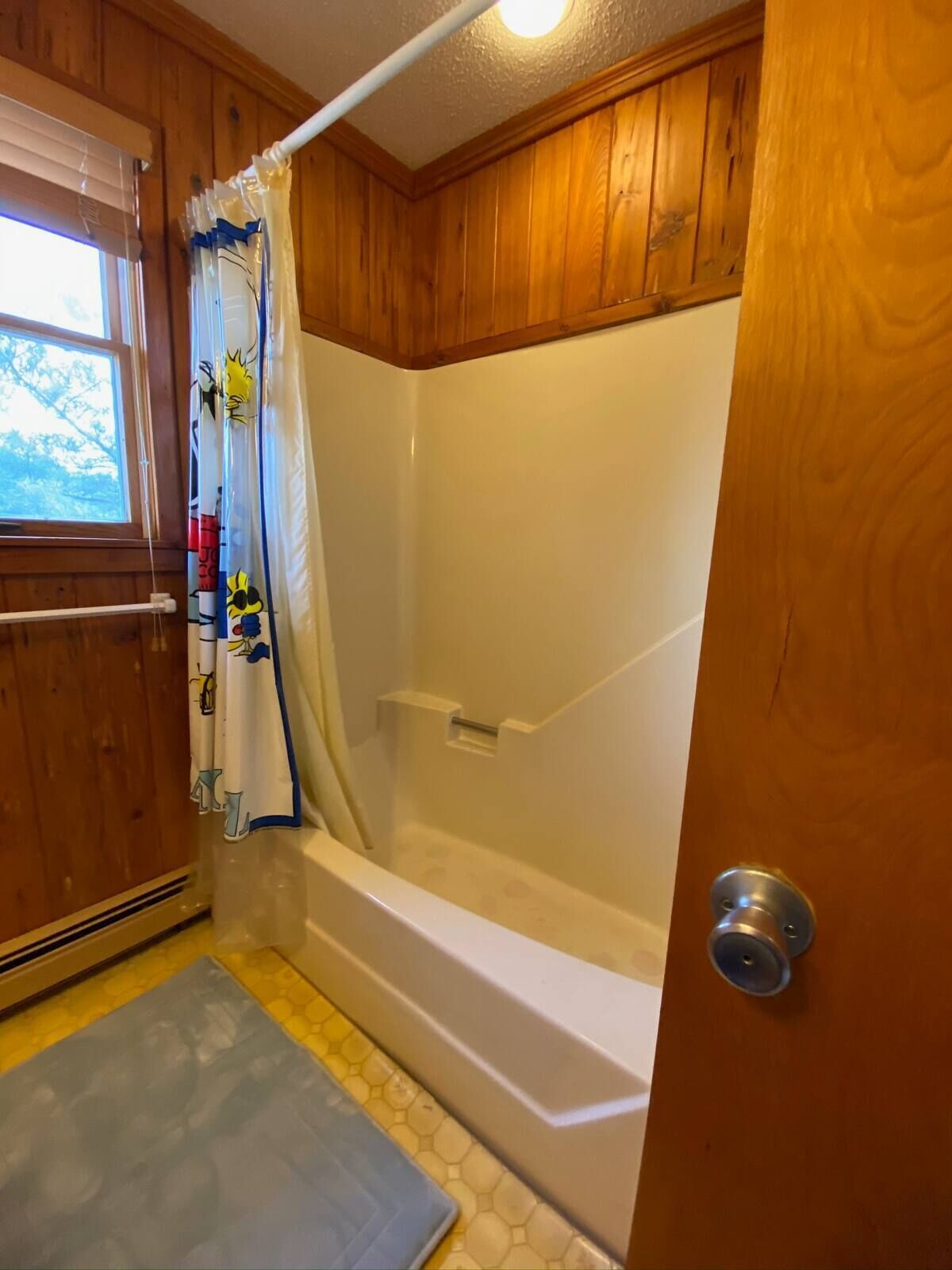 Une salle de bain avec des lambris en bois, une baignoire, un rideau de douche orné de personnages de dessins animés et une fenêtre avec des stores.