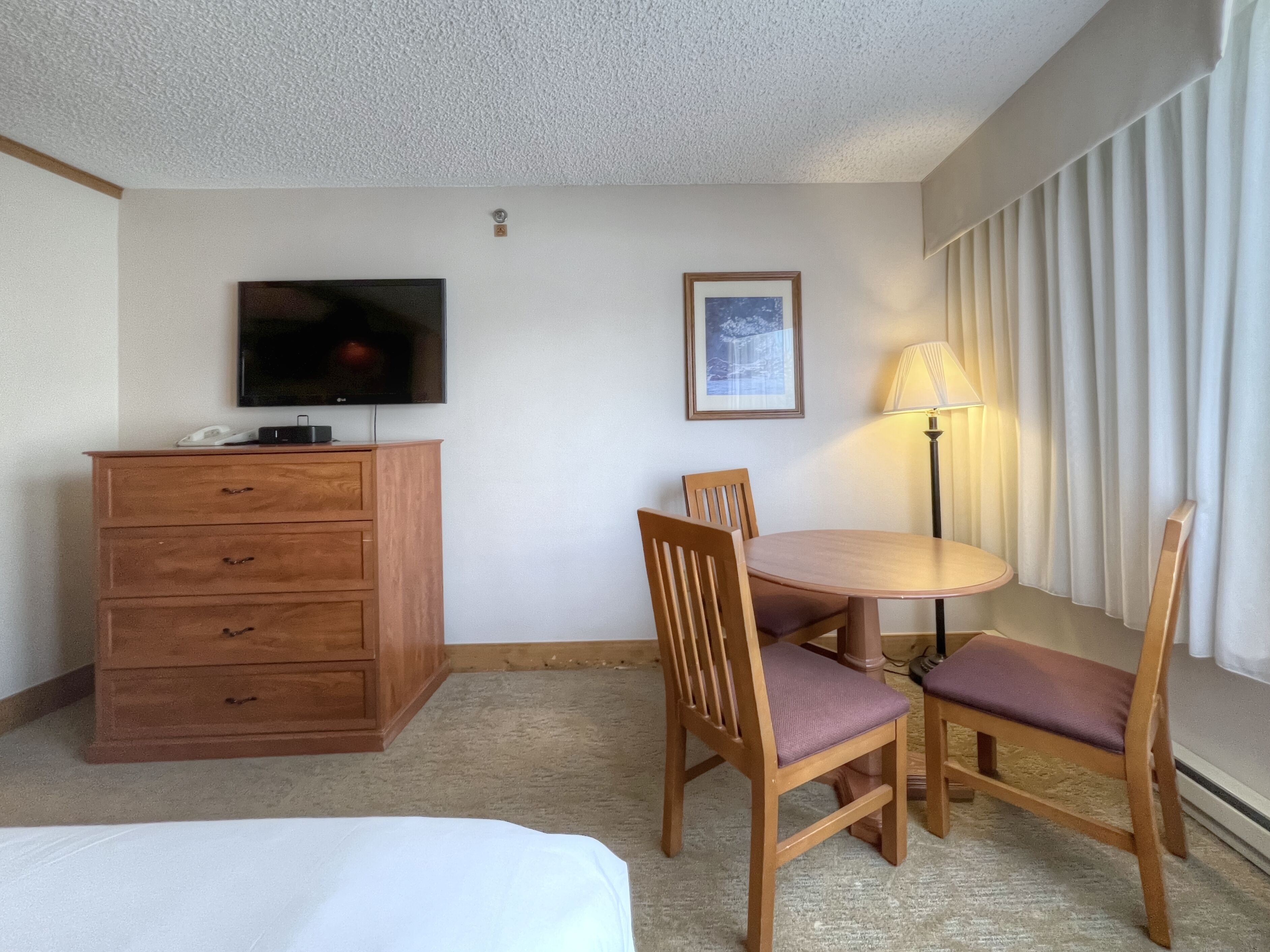 Una habitación de hotel con una cama, un tocador de madera, un televisor, un cuadro enmarco, una mesa redonda con sillas y una lámpara.