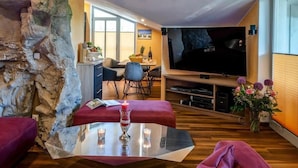 Una sala de estar moderna con una pared de acento de piedra, una mesa de centro de vidrio, asientos rojos, un televisor de pantalla plana y un comedor con sillas y una mesita.