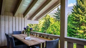 Un espacio de comedor al aire libre cubierto, con mesa de madera y sillas de mimbre, con vista a un jardín exuberante y montañas boscosas.