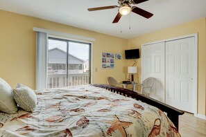 A bedroom with a bed, a ceiling fan, a window with a view of a house, a TV on the wall, and a door. - Reefs IV 1D - Oceanfront Condo in Carolina Beach (Carolina Beach)