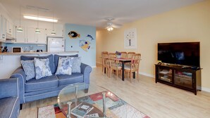Ein Wohnzimmer mit blauem Sofa, einem Glastisch, einer Essecke mit Holzkästen, einem Flachbildfernseher und einer Küche mit weißen Schränken und Pendelleuchten.