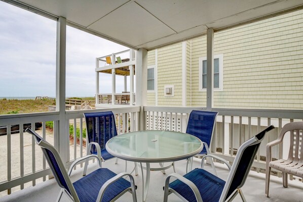 A covered outdoor area with a glass table and four chairs, overlooking a beach.
