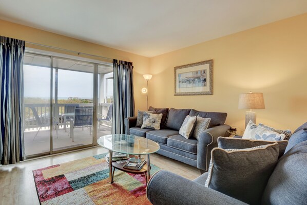 A living room with a sofa, a glass coffee table, and a view through a sliding glass door to an outdoor patio. - Reefs IV 1D - Oceanfront Condo in Carolina Beach (Carolina Beach)