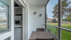 Un balcón con una mesa y sillas de mimbre, una pequeña cocina con microondas y refrigerador, y vista a una zona residencial.
