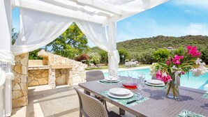 An outdoor patio with a stone fireplace, a dining table set for four, and a view of a swimming pool and hills in the background. - Luxury Villa Laura - new infinity pool, relax and privacy for your holidays (Alghero)