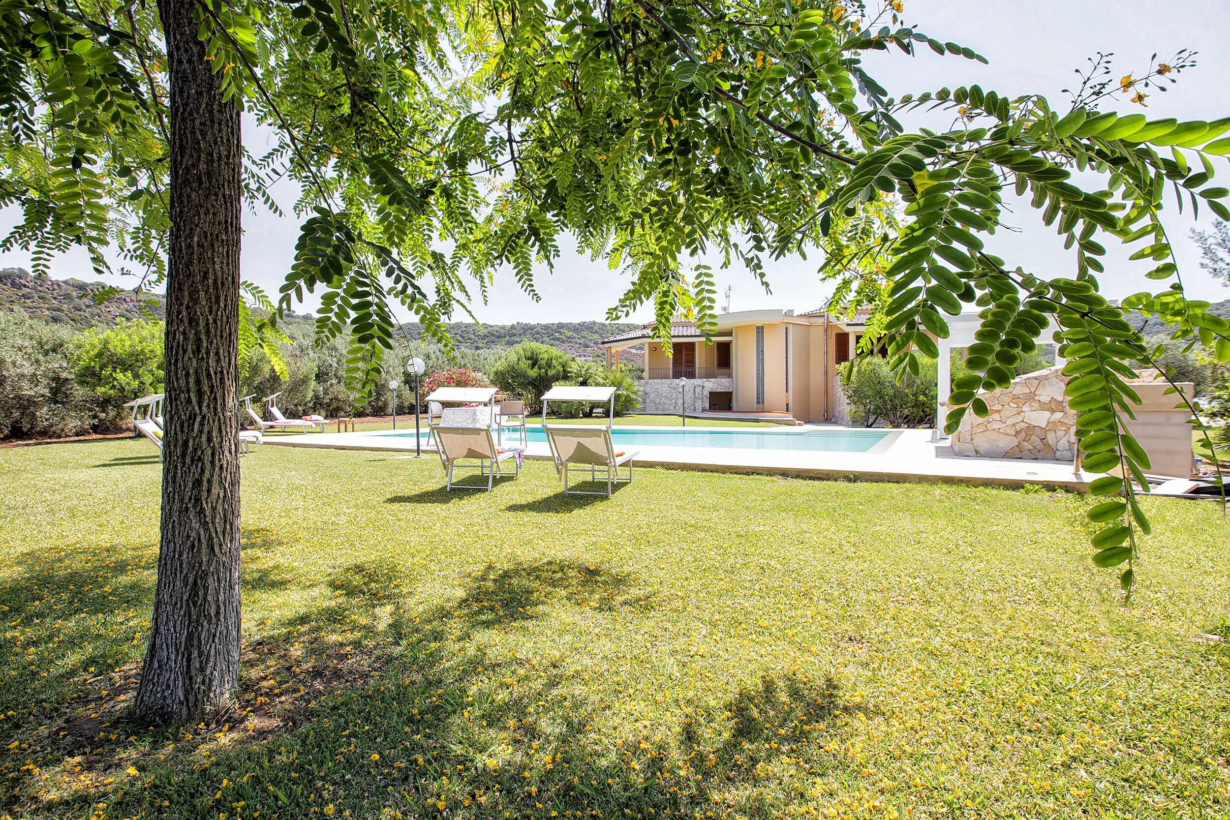 A pool area with lounge chairs, a stone building, and a tree in the foreground.