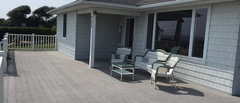 A single-story house with a covered porch, a white fence, and a row of three wicker chairs.