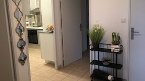 A hallway with a tiled floor, a door leading to a kitchen, and a small shelf with a plant and a vase.