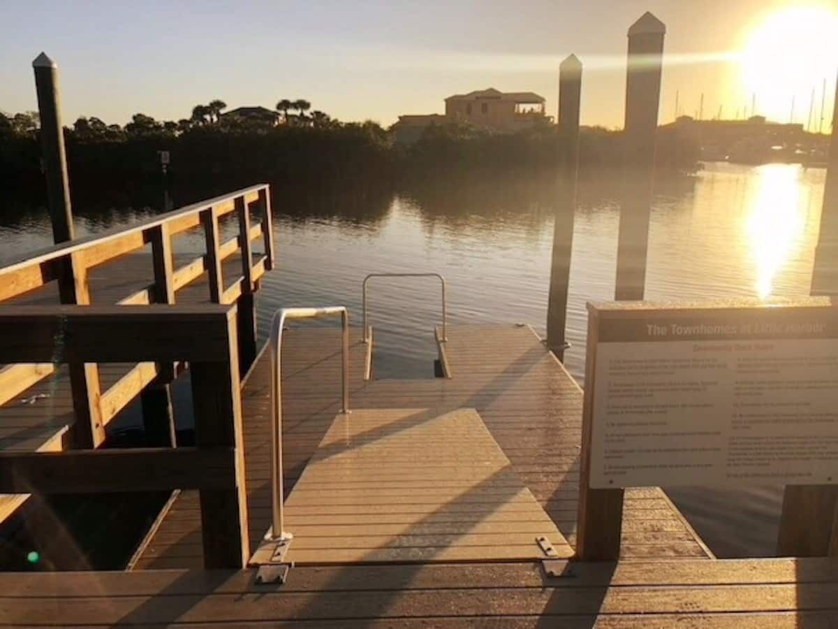 Une jetée en bois avec une rampe menant à une marina, un panneau indicateur et un coucher de soleil en arrière-plan.