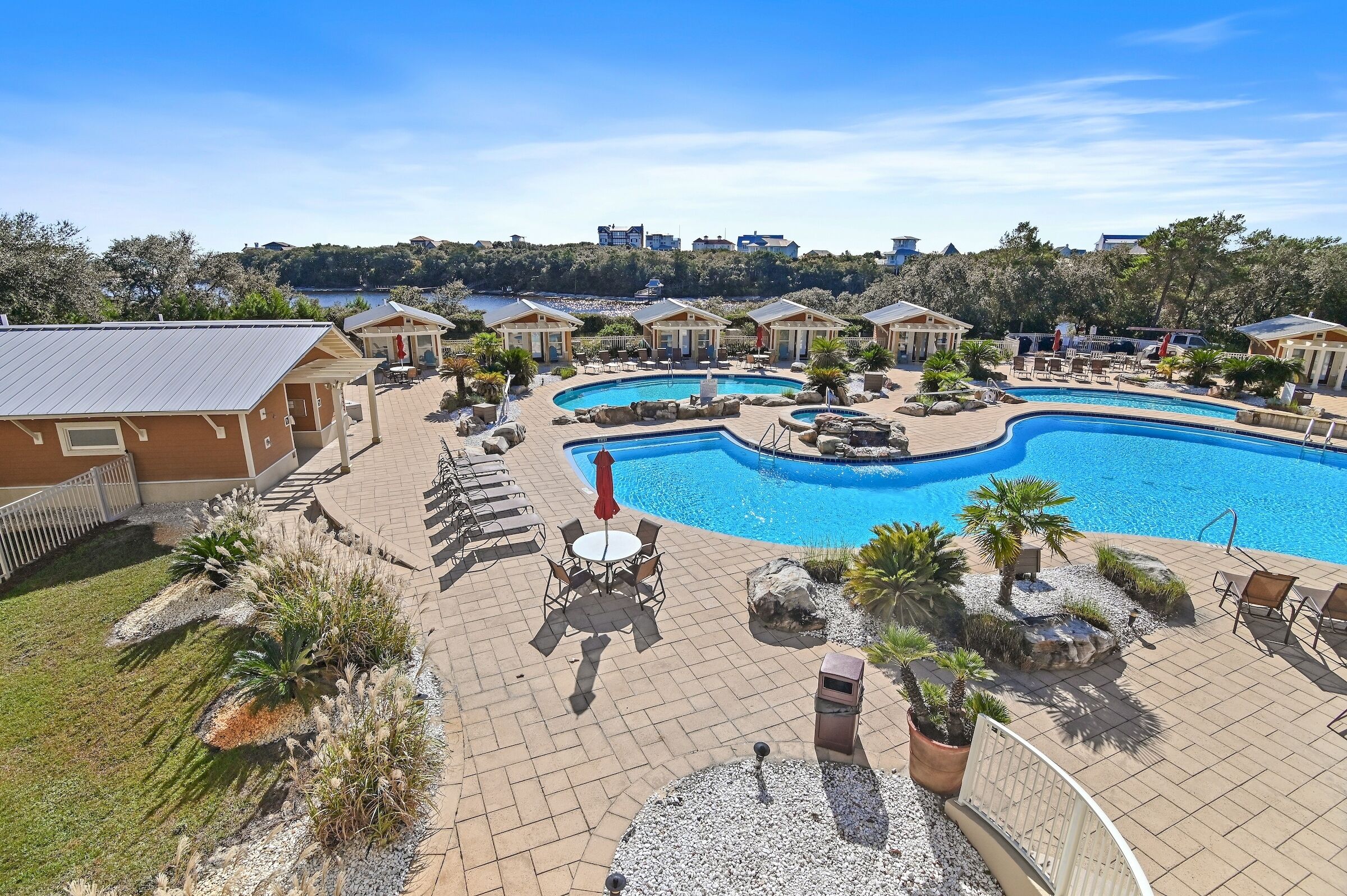 A pool area with lounge chairs and umbrellas, surrounded by small buildings and a view of a hill with buildings in the background.