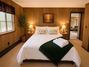 A bedroom with a large bed, two bedside tables with lamps, a painting on the wall, and a view into another room.