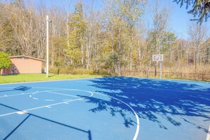 An outdoor basketball court with a blue surface, surrounded by trees and a small building.