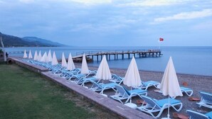 A beach with rows of sun loungers and umbrellas, a pier extending into the water, and a flag indicating the location. - Calidus Hotel (Ayvacik)
