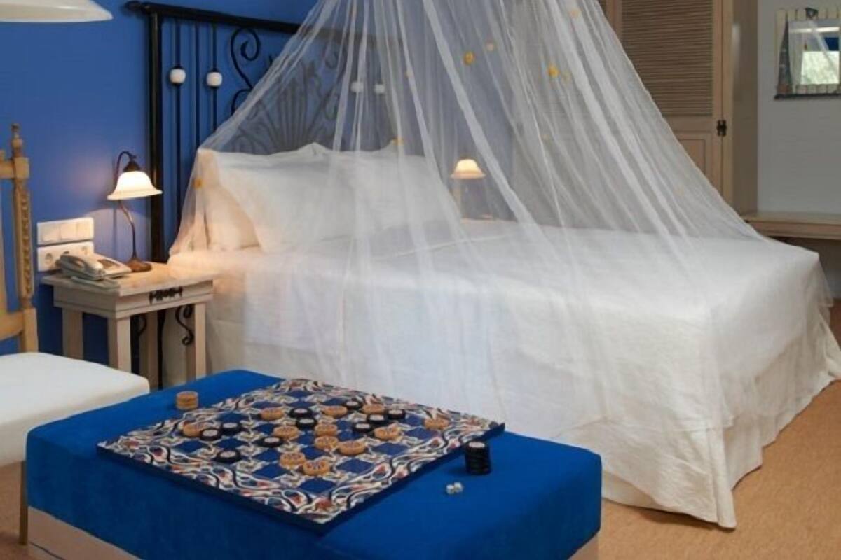 A bedroom with a bed covered by a white canopy, a bedside table with a lamp, and a small table with a colorful mat.