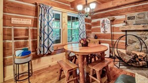 A cabin interior with a wooden dining table, benches, a chandelier, a stone fireplace, and a window with patterned curtains. - Cherry Grove - Rustic Log Cabin (Cherry Creek)