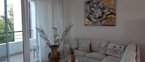 A modern living room with a glass coffee table, a beige sofa, and a large window with a view of greenery.