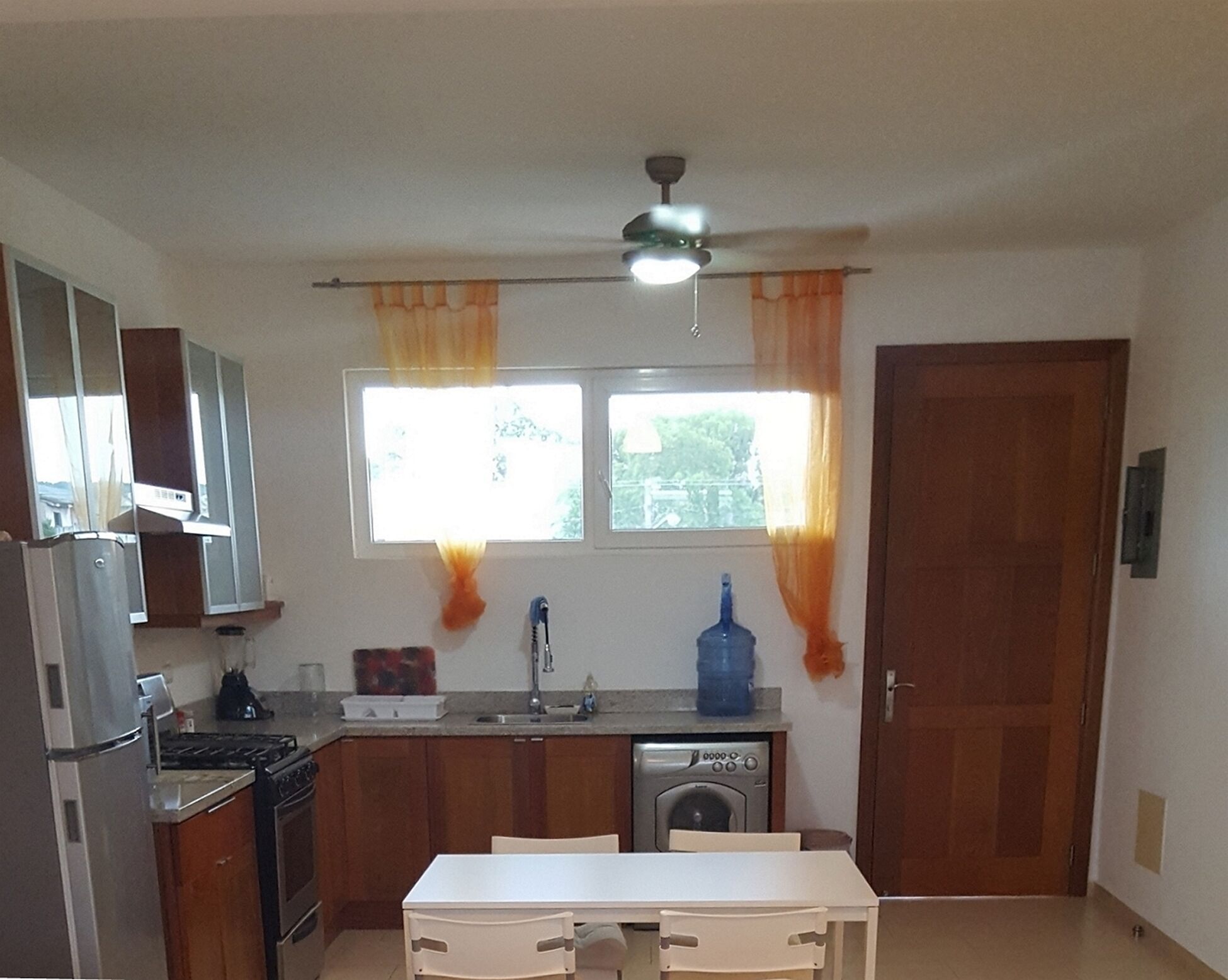 A compact kitchen with a refrigerator, stove, sink, and dishwasher. There is a window with curtains, a ceiling fan, and a door.