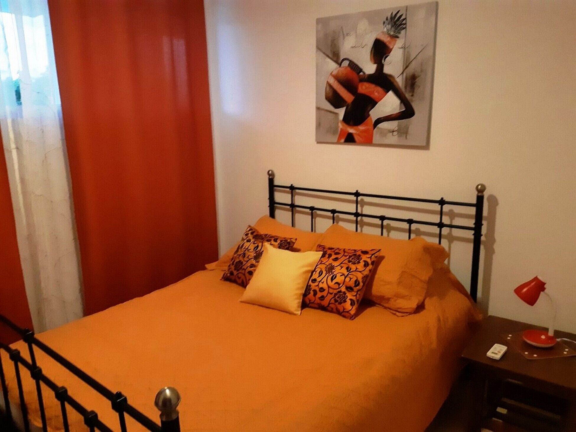A bed with an orange bedspread and patterned pillows, a black metal headboard, and a red lamp on a bedside table.