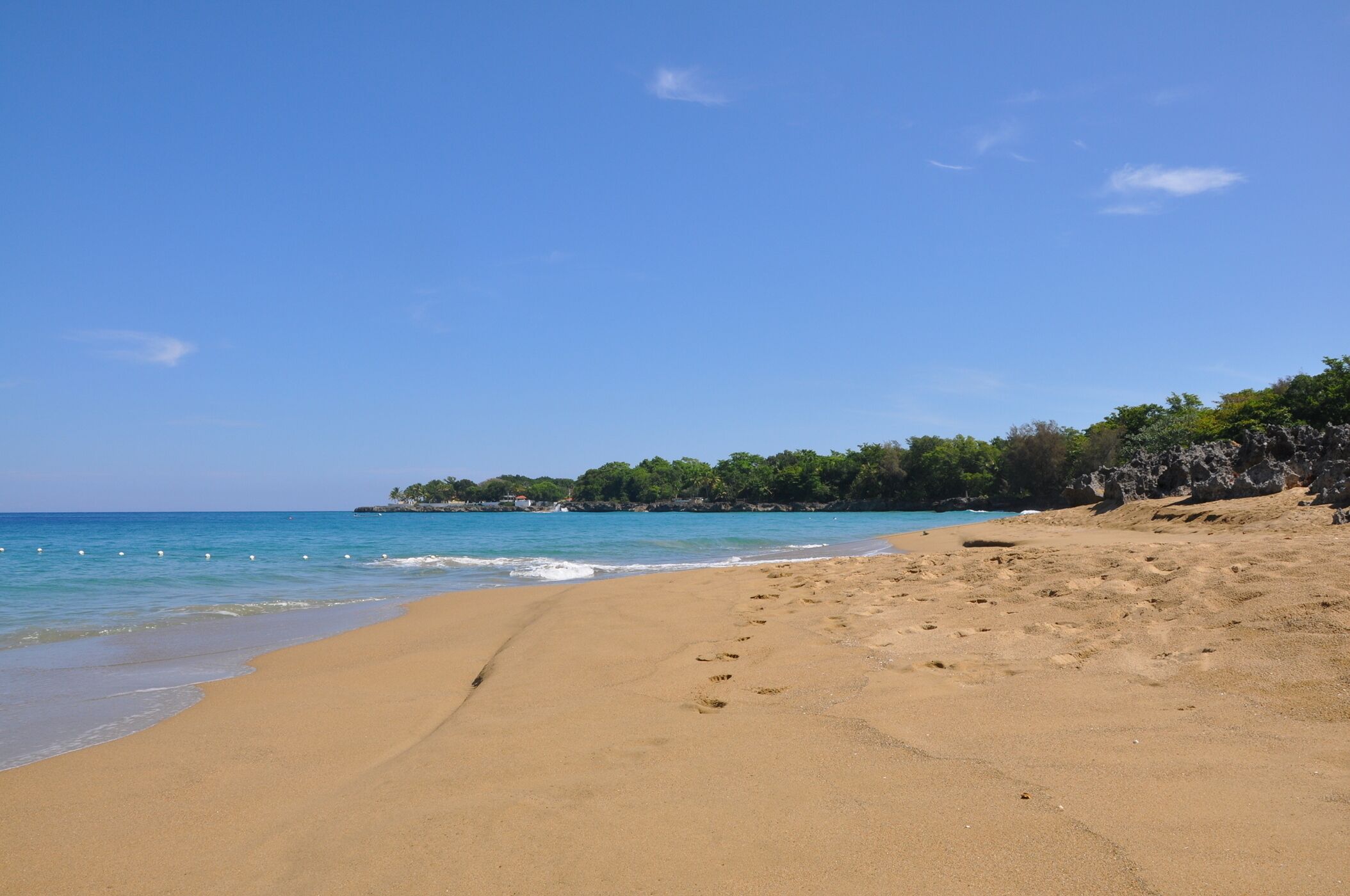 A beach with clear blue water, sandy shore, and a rocky outcrop with vegetation.
