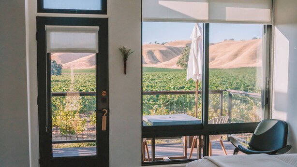 A bedroom with a large bed, a view of a vineyard through a sliding door, and a balcony with a table and chairs. - Geneseo Inn (Paso Robles)