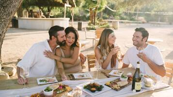 Un grupo de personas cenando en una mesa al aire libre con diferentes comidas y bebidas.