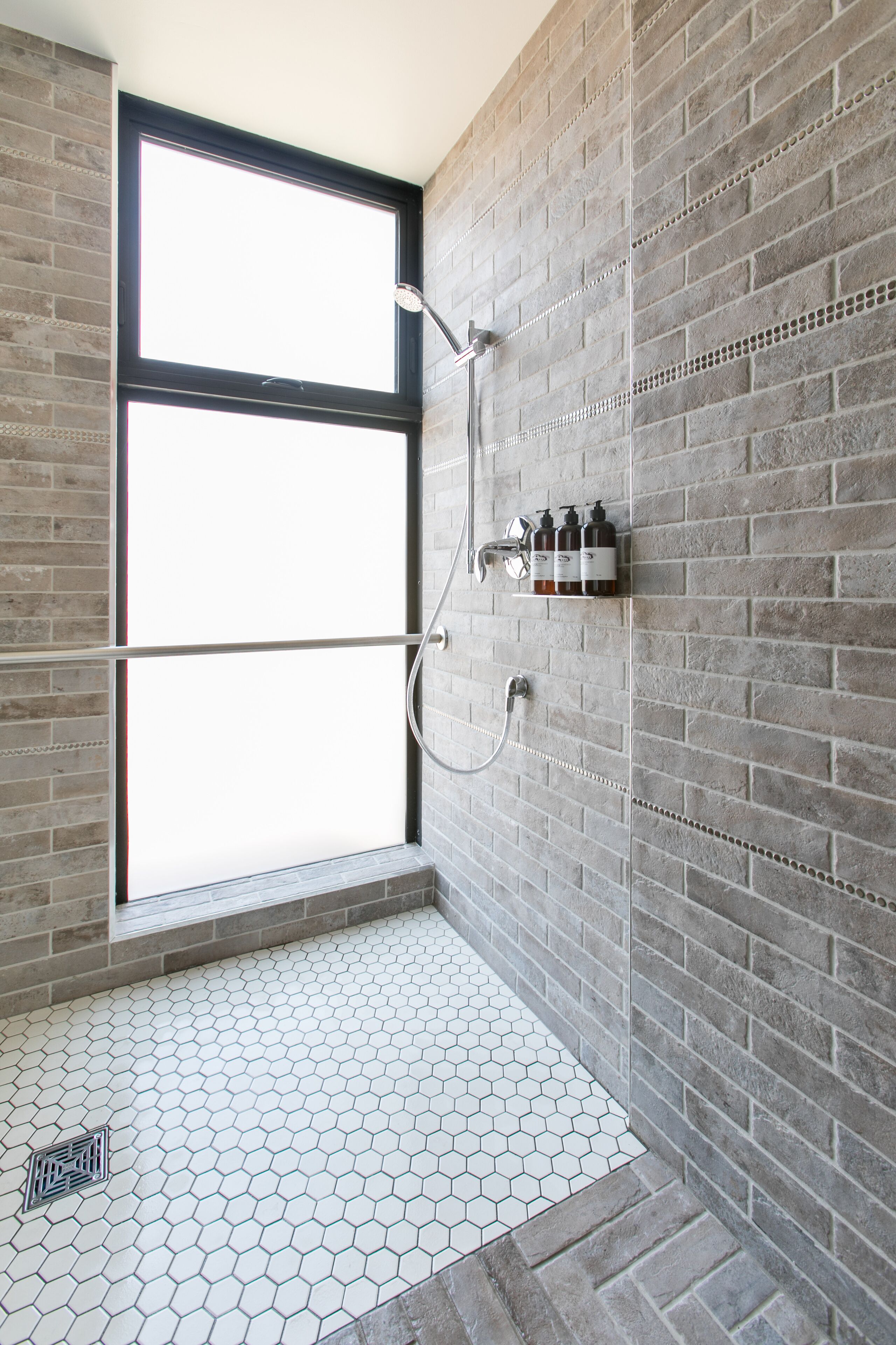 A modern bathroom with a large window, a shower with a handheld showerhead, and a shelf with toiletries.
