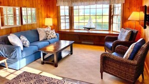 Ein gemütliches Wohnzimmer mit einem blauen Sofa, einem Holztisch, Korbstühlen und einem großen Fenster mit Blick auf Grünflächen.