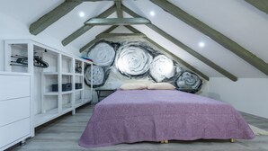 Une chambre avec un plafond en pente, un lit recouvert d’une courtepointe violette, une armoire blanche et un grand tableau abstrait accroché au mur.