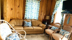 A cozy living room with a brown leather sofa, two wicker chairs, and a television on a wooden wall. - Waterfront Cabin #1, Onalaska, Lake Livingston, Texas (Onalaska)