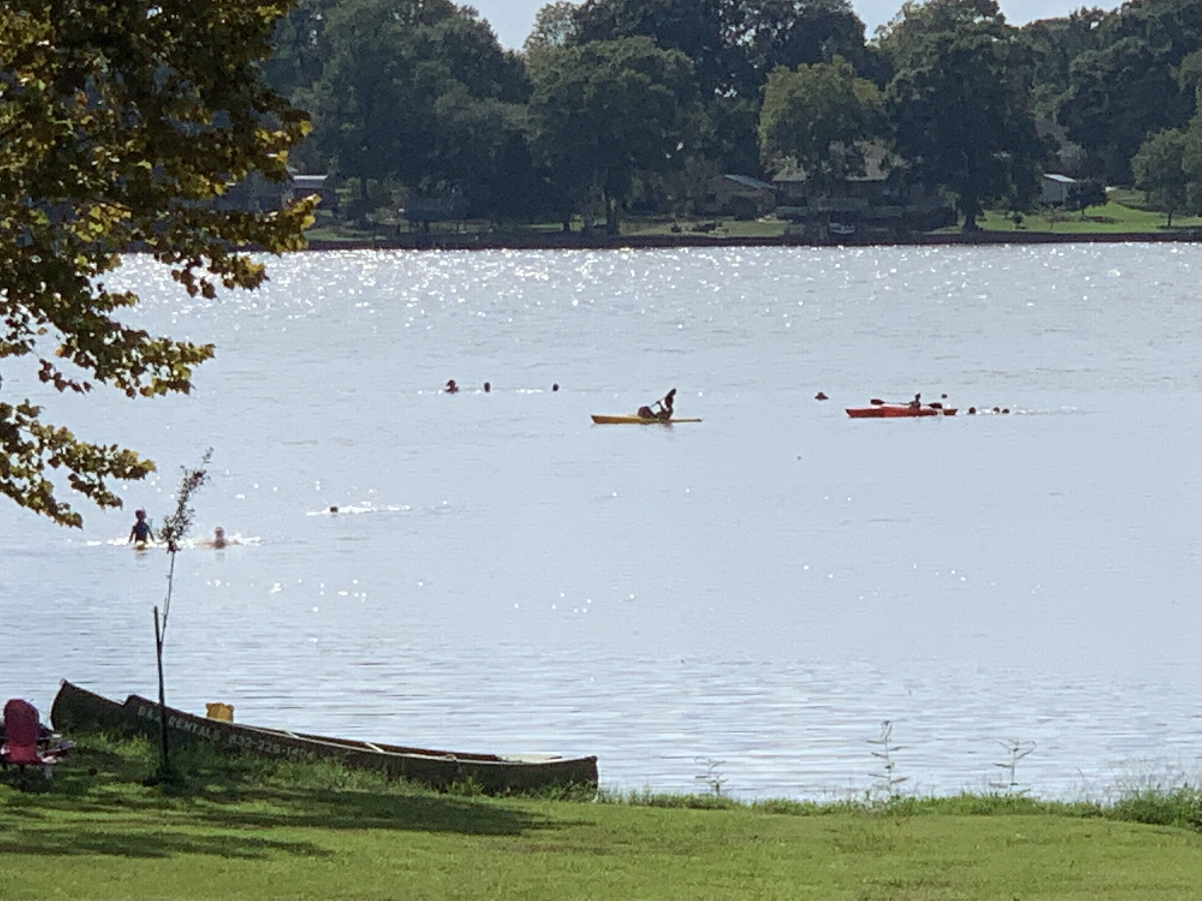 A calm lake with people kayaking and canoeing. There is a dock with a boat and a small structure on the shore.