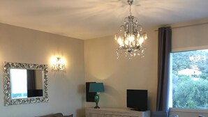 A modern living room with a grey sofa, a dining table set for four, a chandelier, a large mirror, and a TV.