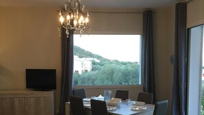 A dining area with a large table, chairs, a chandelier, a television on a cabinet, and a view of greenery and a building through large windows.