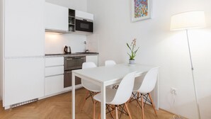 Una cucina compatta con mobili bianchi, un forno a microonde e un piano cottura. C'è un tavolo da pranzo con quattro sedie e una pianta in vaso. Una lampada da terra si trova sulla destra.