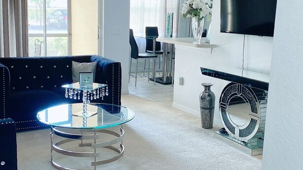 A spacious living room with a round glass table, a dark blue sofa, a chandelier, a flat-screen TV, and a dining area with chairs.