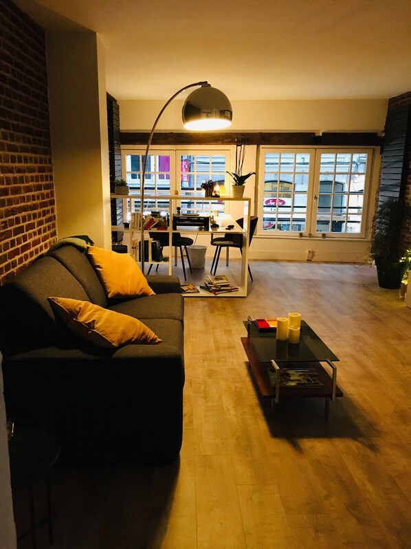 Una sala moderna con un sofá de color oscuro, una mesita de centro y una lámpara de pie. Se ve un comedor con una mesa y sillas al fondo.