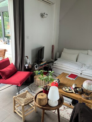 A modern living room with a red chair, a wooden coffee table, a TV, and a bed with white bedding.