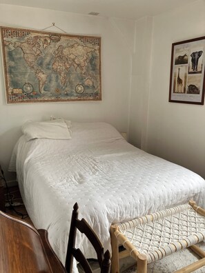 A neatly made bed with a white duvet, a wooden chair with a woven seat, a world map on the wall, and a framed picture of African animals.