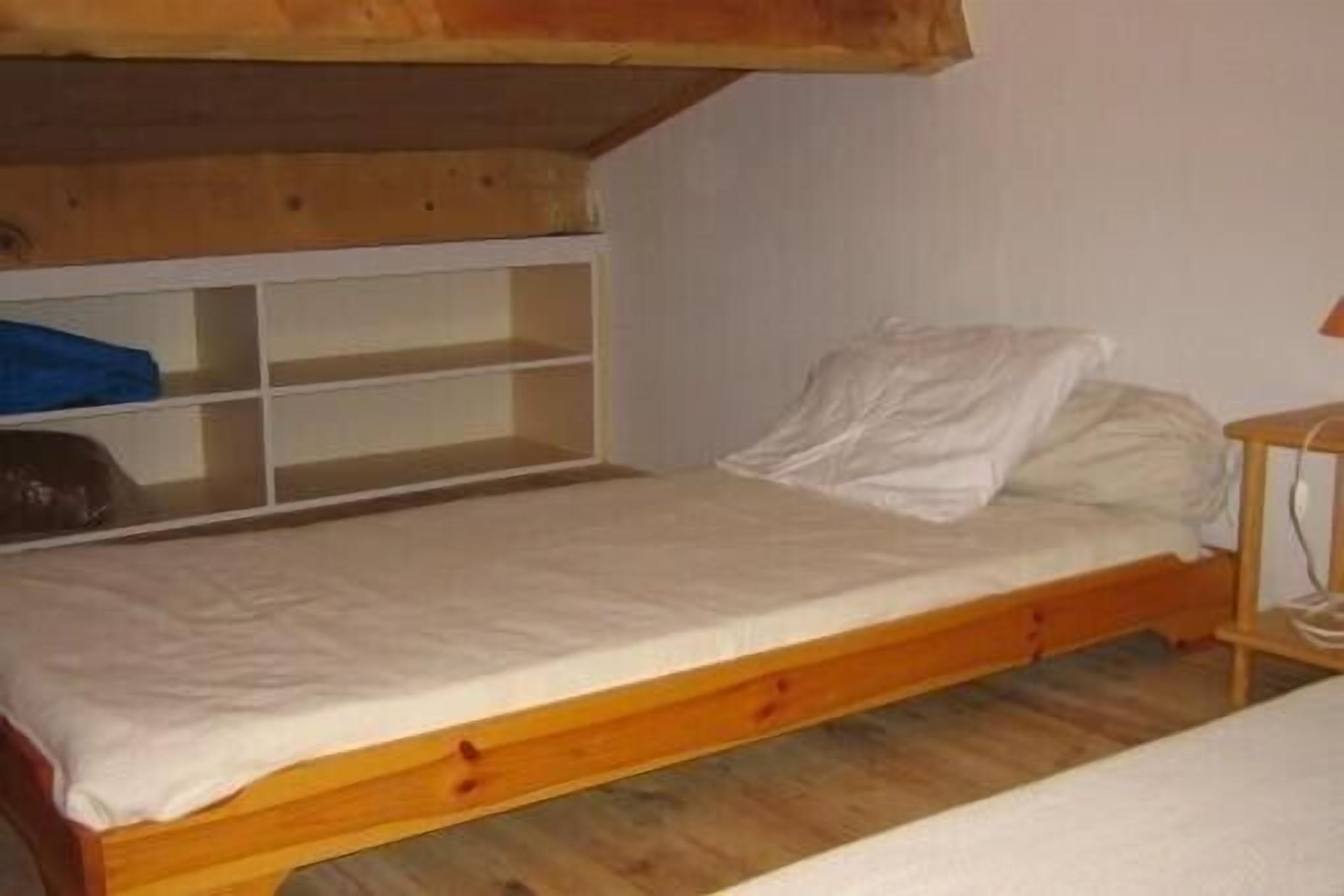 Ein Einzelbett mit Holzrahmen und weißer Matratze, darüber ein Holzregal und ein kleiner Nachttisch mit Lampe.