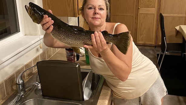 A person holding a large fish in a kitchen with wooden cabinets, a sink, and a window with blinds.