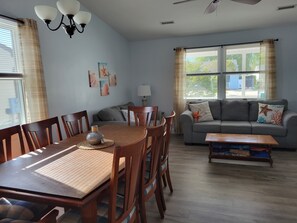 Un salon comprenant une table à manger, des chaises, un canapé et une table basse. On y trouve des fenêtres avec des rideaux, un ventilateur de plafond et des œuvres d’art murales.